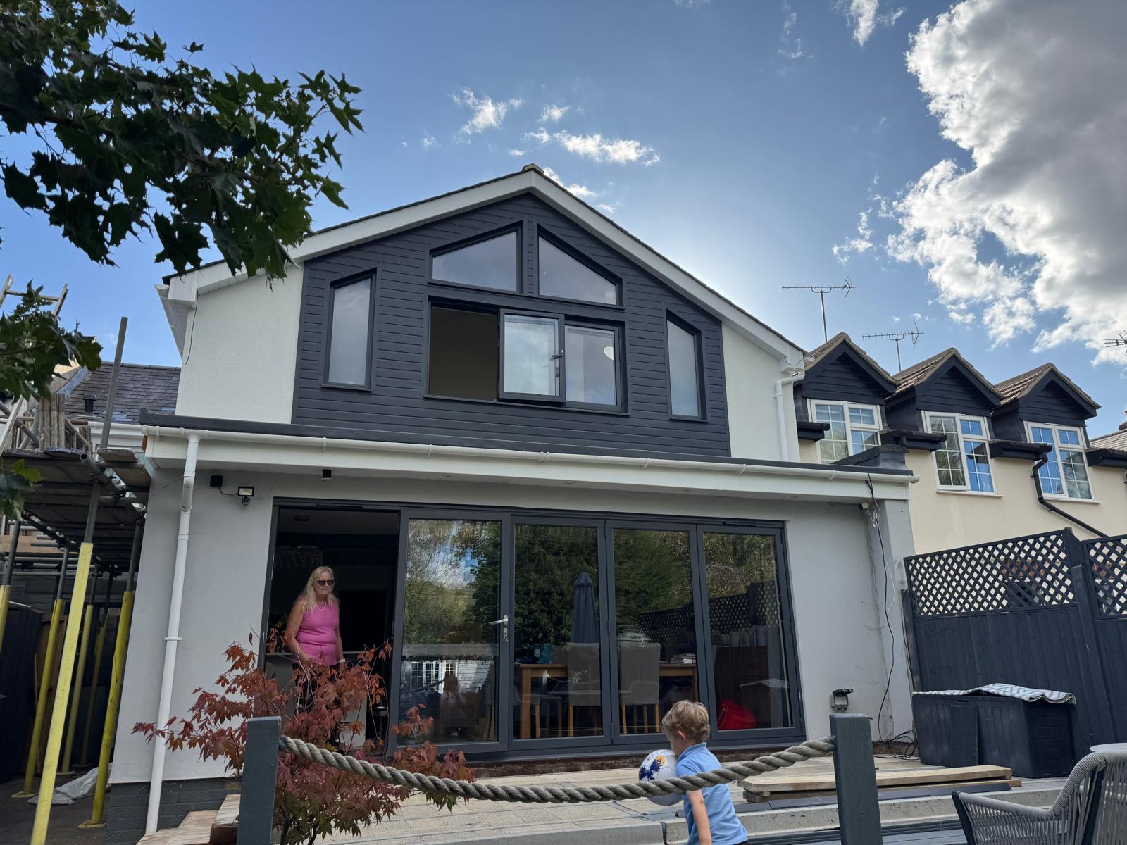 Modern two-storey residential home extension featuring dark composite cladding, bespoke gable windows, and expansive bi-fold doors opening to a paved patio.